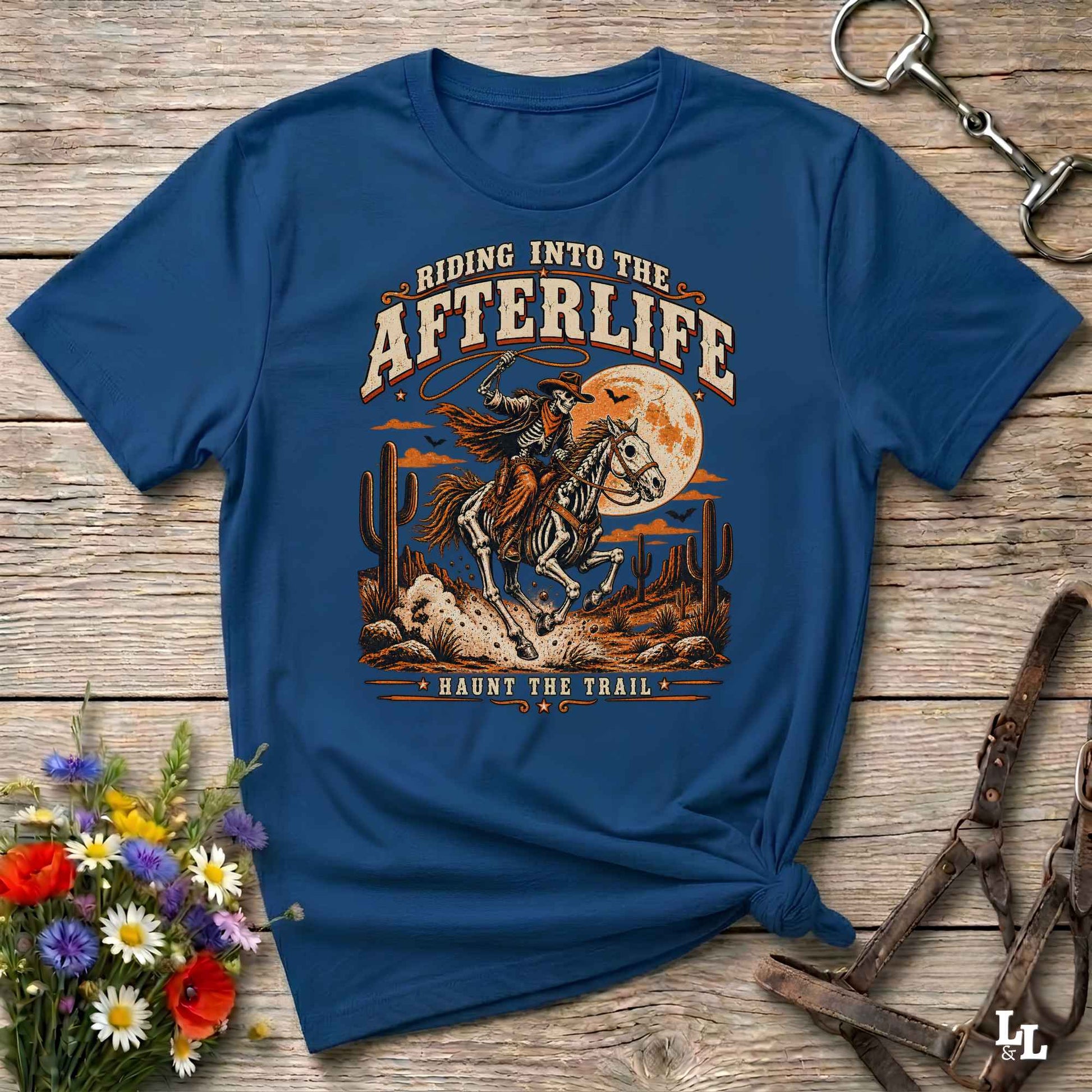 A navy blue graphic t-shirt featuring a western-themed design of a cowboy riding a horse across a desert landscape with a full moon. The text reads 'RIDING INTO THE AFTERLIFE' and 'HAUNT THE TRAIL'.