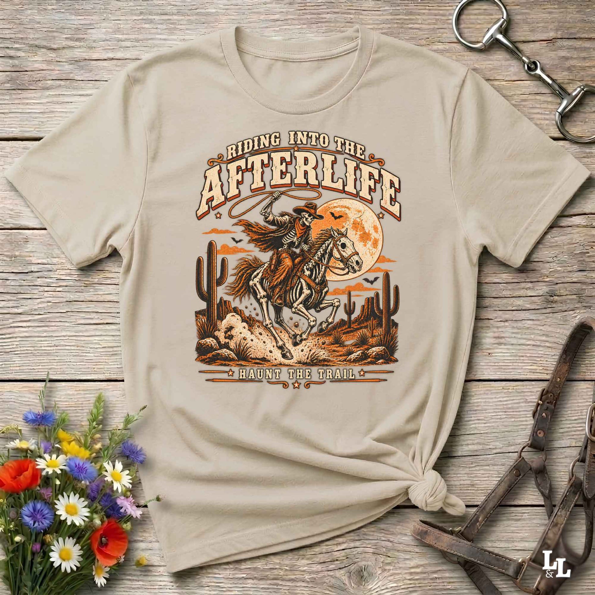 A beige graphic t-shirt with a western design, laid flat on a rustic wooden background with decorative props including a horse bit, leather reins, and a small bouquet of flowers.