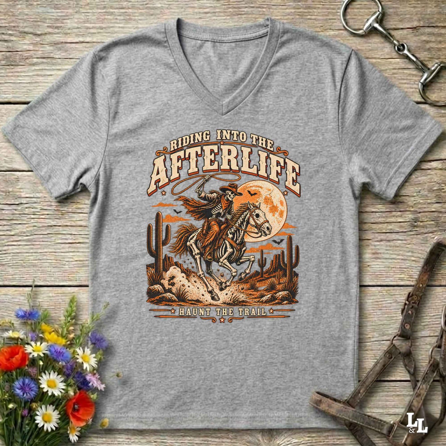 A heather gray V-neck t-shirt with a western-themed graphic print reading 'Riding Into the Afterlife' and 'Follow the Trails,' featuring a cowboy on a horse in a desert landscape with cacti and a full moon, laid flat on a rustic wood background with flowers and horse tack props.