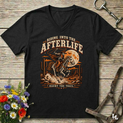 A black V-neck t-shirt with a retro-style graphic of a skeleton cowboy riding a horse under a full moon, with the text 'RIDING INTO THE AFTERLIFE' and 'HAUNTS THE TRAIL'. The shirt is displayed flat on a rustic wooden background with horse tack, flowers, and a bit of grass.