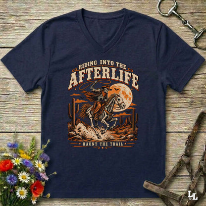 A navy blue V-neck t-shirt with a vintage-style western graphic of a cowboy riding a horse under a full moon, with the text 'RIDING INTO THE AFTERLIFE' and 'HAUNT THE TRAIL'. The shirt is displayed flat on a rustic wooden background with equestrian accessories and flowers.