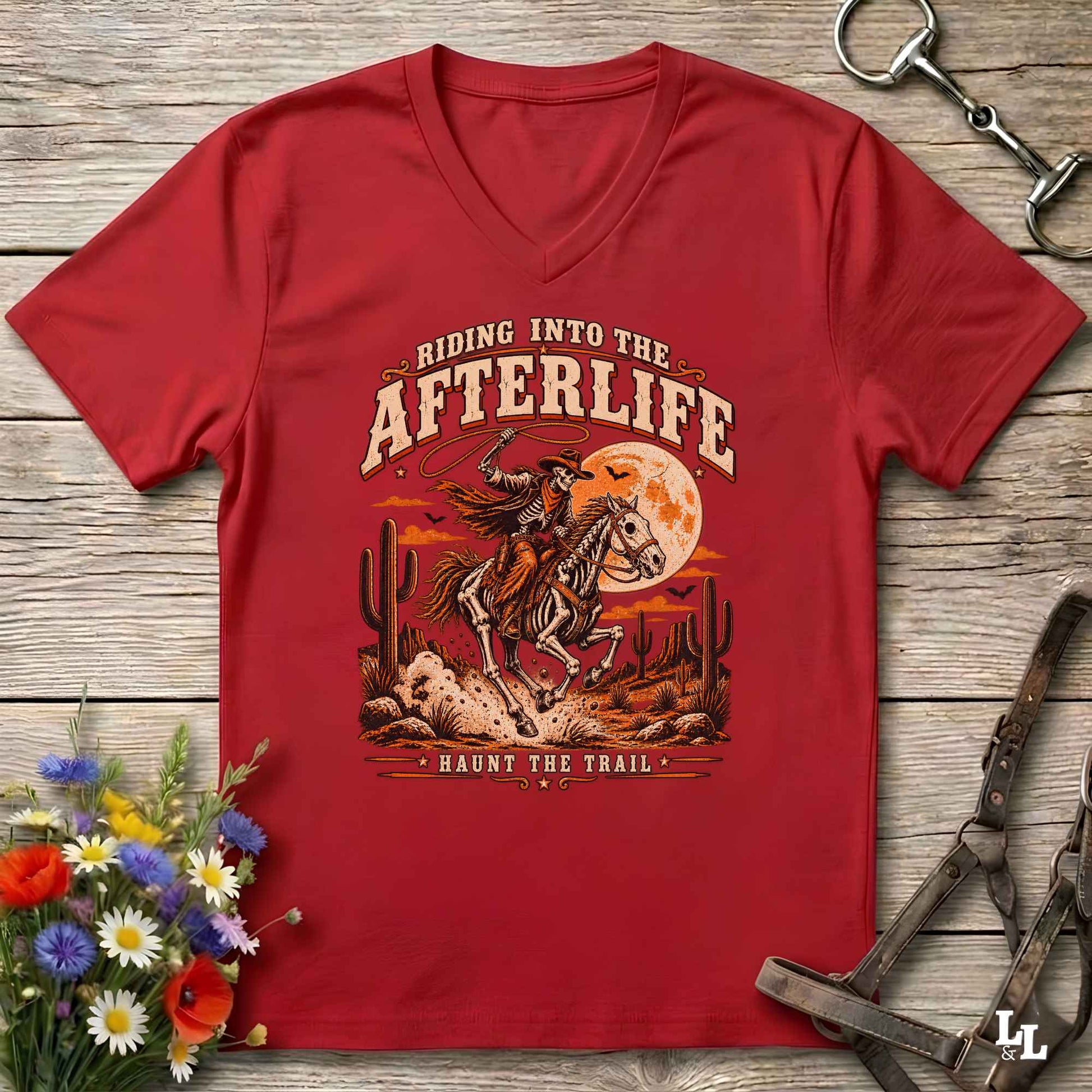 A red V-neck t-shirt laid flat on a rustic wooden surface, featuring a western-style graphic of a skeleton cowboy riding a horse under a full moon with the text 'Riding Into the Afterlife' and 'Haunt the Trail.' Decorative flowers and horse tack are placed around the shirt.
