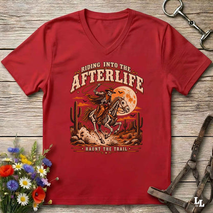A red V-neck t-shirt laid flat on a rustic wooden surface, featuring a western-style graphic of a skeleton cowboy riding a horse under a full moon with the text 'Riding Into the Afterlife' and 'Haunt the Trail.' Decorative flowers and horse tack are placed around the shirt.