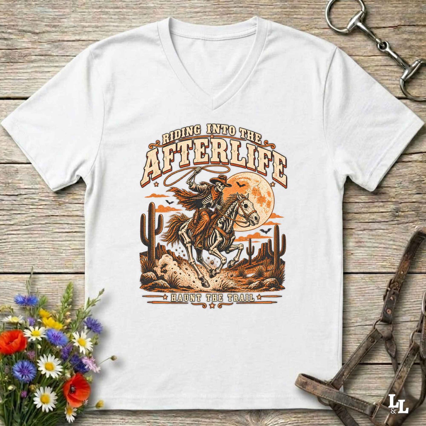 A white V-neck t-shirt with a western-themed graphic print reading 'Riding Into the AFTERLIFE' and 'Have No The Trail', featuring an illustration of a cowboy on a horse with cacti and an orange moon, displayed on a rustic wooden background with decorative props.