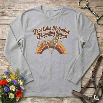 A light gray long-sleeve t-shirt with a retro graphic of a skeleton horse under a rainbow and the text 'Trot Like Nobody's Haunting You'. The shirt is displayed flat on a rustic wooden background with decorative props including a bouquet of flowers, a leather bridle, and a horse bit.