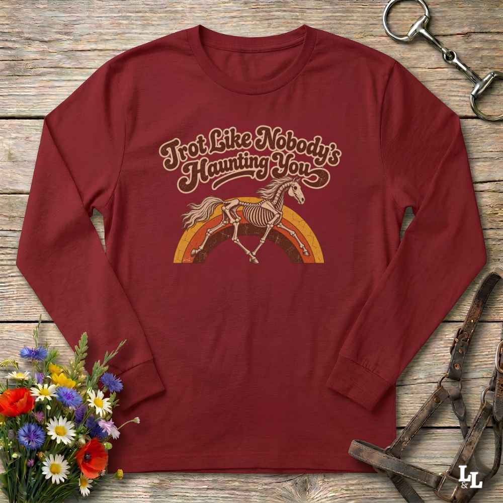 A long-sleeve crewneck shirt in maroon red, laid flat on a rustic wooden surface. The shirt features a graphic print of a skeleton horse running under a rainbow with the text 'I'm Not Like Nobody's Haunting You' in a retro font.