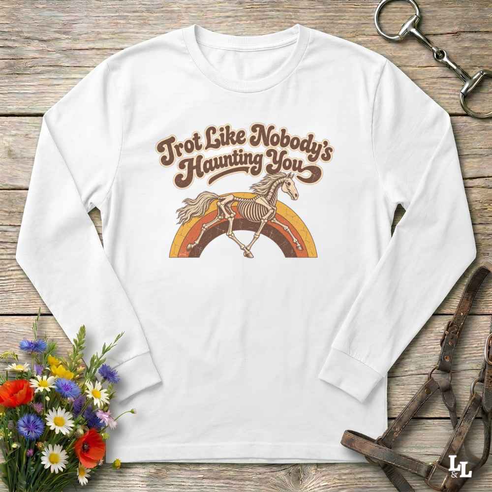 A white long-sleeve t-shirt with a retro-style graphic print featuring the text 'Trot Like Nobody's Haunting You' above a skeleton horse running over a rainbow. The shirt is displayed in a flat-lay arrangement on a rustic wooden background, styled with equestrian leather tack and a small bouquet of wildflowers.