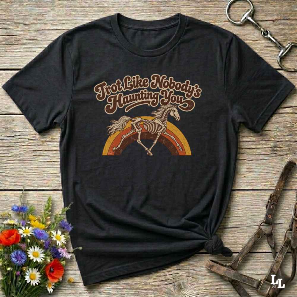 A black short-sleeve graphic T-shirt laid flat on a rustic wooden surface, styled with flowers, a horse bit, and leather tack. The shirt features a retro-style printed design with the phrase 'I'm Not Like Nobody's Haunting You' above an illustration of a horse skeleton running over a rainbow.