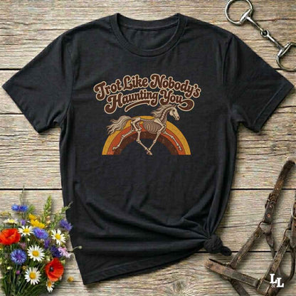 A black short-sleeve graphic T-shirt laid flat on a rustic wooden surface, styled with flowers, a horse bit, and leather tack. The shirt features a retro-style printed design with the phrase 'I'm Not Like Nobody's Haunting You' above an illustration of a horse skeleton running over a rainbow.