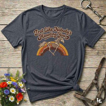 A dark heather gray graphic t-shirt featuring a skeleton horse running over a rainbow with the text 'Trot Like Nobody's Haunting You'. The shirt is styled in a flat-lay on a rustic wooden background with decorative elements including a halter, a horse bit, and a small bouquet of wildflowers.