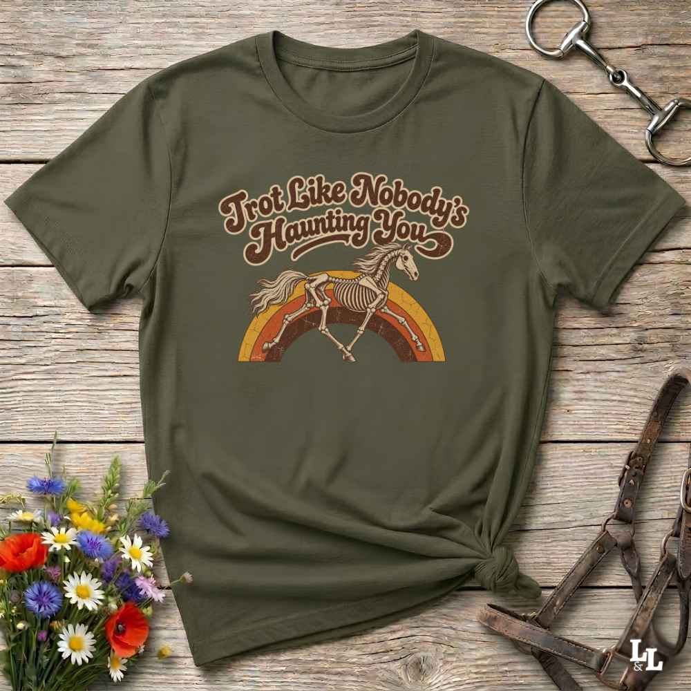 An olive green short-sleeve t-shirt with a retro graphic of a horse skeleton jumping over a rainbow, laid flat on a rustic wooden surface with decorative flowers and leather tack around it.