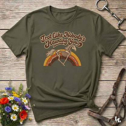 An olive green short-sleeve t-shirt with a retro graphic of a horse skeleton jumping over a rainbow, laid flat on a rustic wooden surface with decorative flowers and leather tack around it.