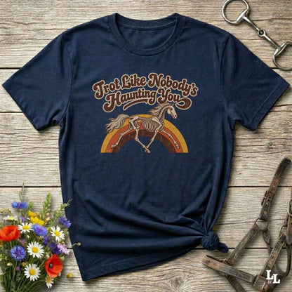 A navy blue short-sleeve t-shirt with a graphic print of a skeleton horse on a rainbow and the text 'Trot Like Nobody's Haunting You'. The shirt is styled flat on a rustic wooden background with decorative flowers and leather tack.