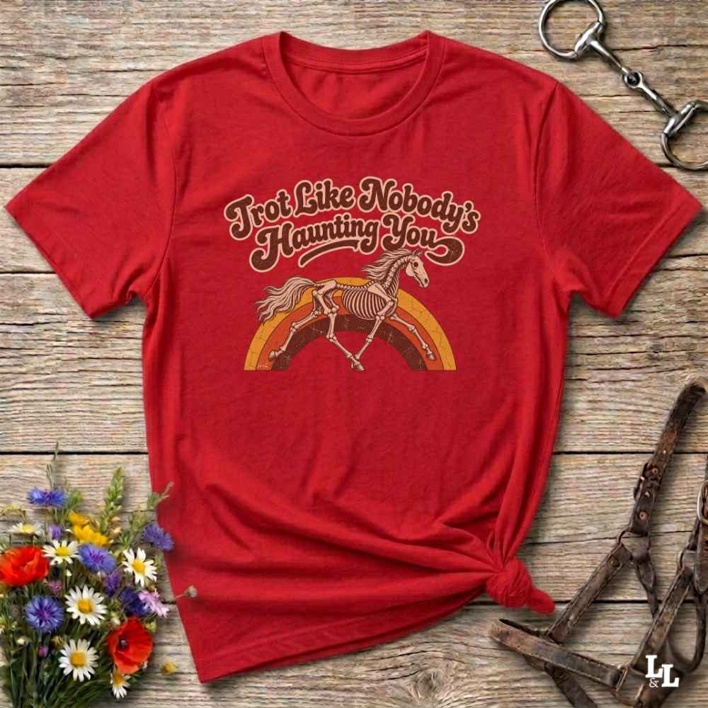 A red short-sleeve graphic T-shirt laid flat on a rustic wooden surface, styled with flowers, a horse bit, and a leather bridle. The shirt has a vintage-style chest graphic with the phrase 'Trot Like Nobody's Haunting You' above a skeleton horse riding a rainbow.