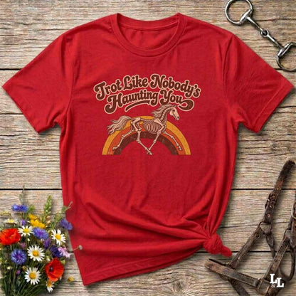 A red short-sleeve graphic T-shirt laid flat on a rustic wooden surface, styled with flowers, a horse bit, and a leather bridle. The shirt has a vintage-style chest graphic with the phrase 'Trot Like Nobody's Haunting You' above a skeleton horse riding a rainbow.