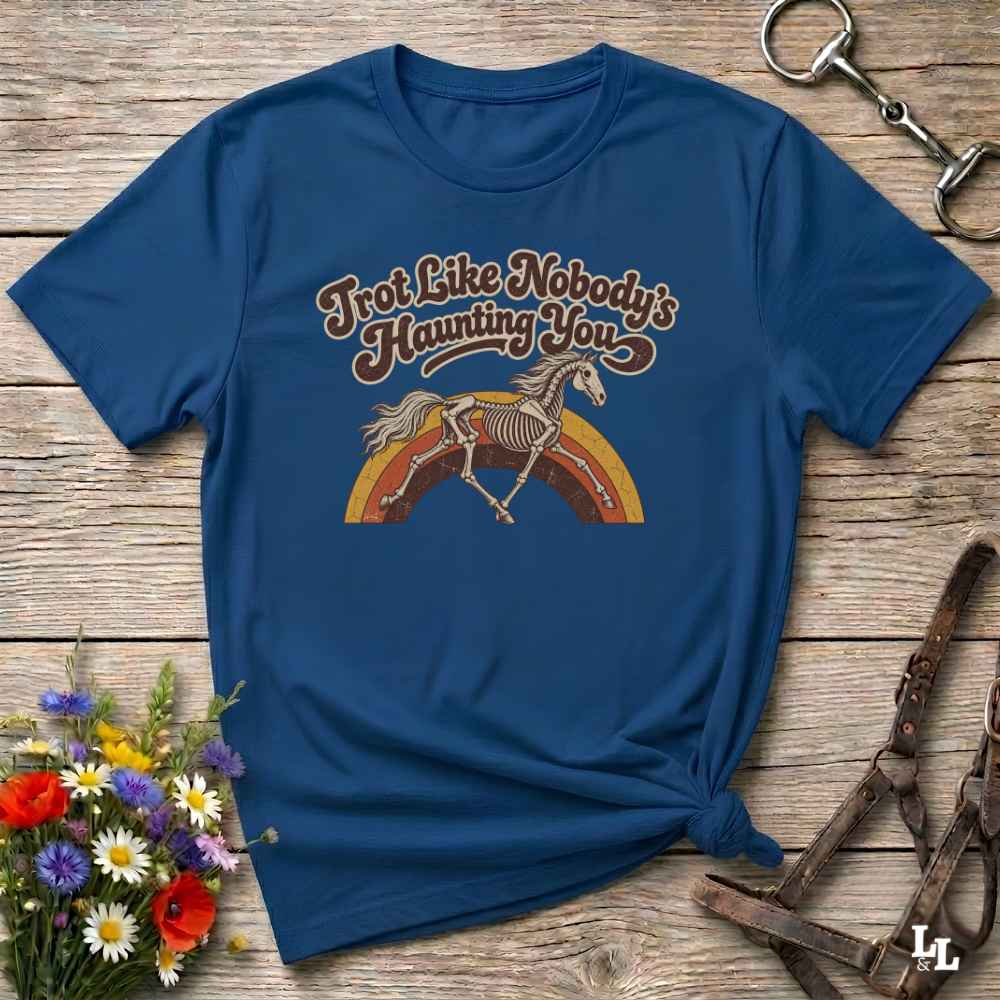 A navy blue short-sleeve t-shirt with a retro-style graphic of a skeleton horse on a rainbow and the text 'Trot Like Nobody's Haunting You'. The shirt is styled in a flat-lay on a rustic wooden background with equestrian accessories and flowers.