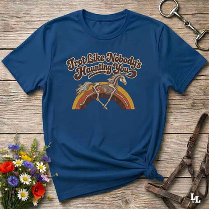 A navy blue short-sleeve t-shirt with a retro-style graphic of a skeleton horse on a rainbow and the text 'Trot Like Nobody's Haunting You'. The shirt is styled in a flat-lay on a rustic wooden background with equestrian accessories and flowers.