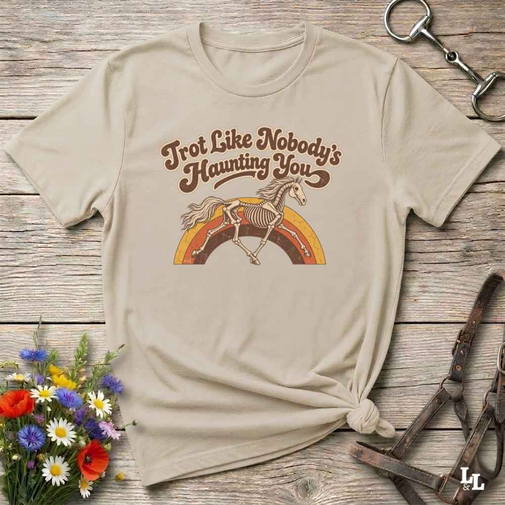 A beige graphic t-shirt laid flat on a rustic wooden surface, styled with equestrian accessories and a small bouquet of wildflowers. The shirt features a retro-style brown graphic of a horse skeleton jumping over a rainbow with the text 'Trot Like Nobody's Haunting You'.