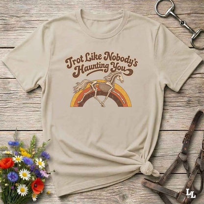 A beige graphic t-shirt laid flat on a rustic wooden surface, styled with equestrian accessories and a small bouquet of wildflowers. The shirt features a retro-style brown graphic of a horse skeleton jumping over a rainbow with the text 'Trot Like Nobody's Haunting You'.