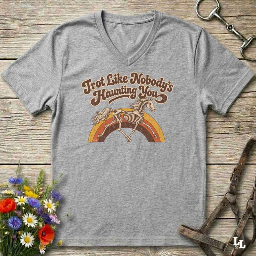 A heather gray V-neck t-shirt with a retro graphic of a skeleton horse running over a rainbow, with the text 'Trot Like Nobody's Haunting You'. The shirt is displayed flat on a rustic wooden background with decorative equestrian and floral props.