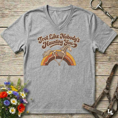 A heather gray V-neck t-shirt with a retro graphic of a skeleton horse running over a rainbow, with the text 'Trot Like Nobody's Haunting You'. The shirt is displayed flat on a rustic wooden background with decorative equestrian and floral props.