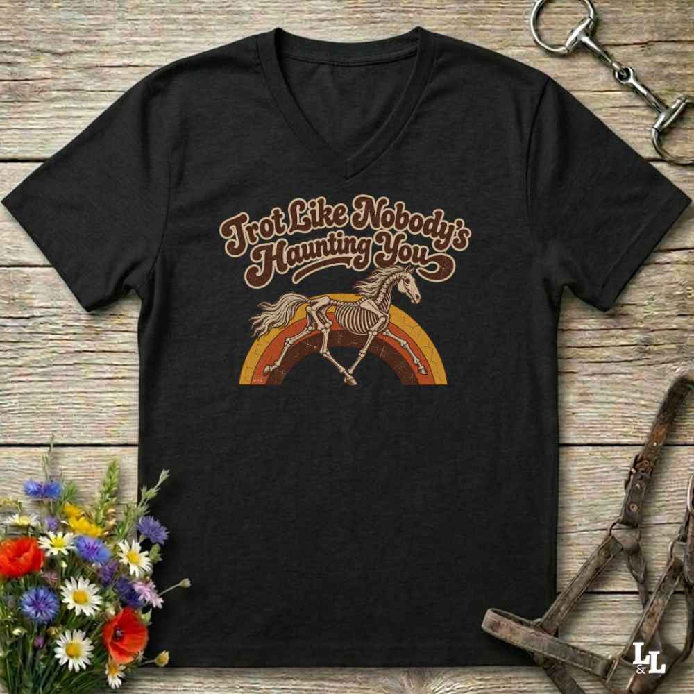 A black V-neck t-shirt with a retro graphic of a horse skeleton over a rainbow and the text 'I'm Not Like Nobody's Haunting You', displayed on a rustic wooden background with equestrian accessories and flowers.