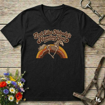 A black V-neck t-shirt with a retro graphic of a horse skeleton over a rainbow and the text 'I'm Not Like Nobody's Haunting You', displayed on a rustic wooden background with equestrian accessories and flowers.
