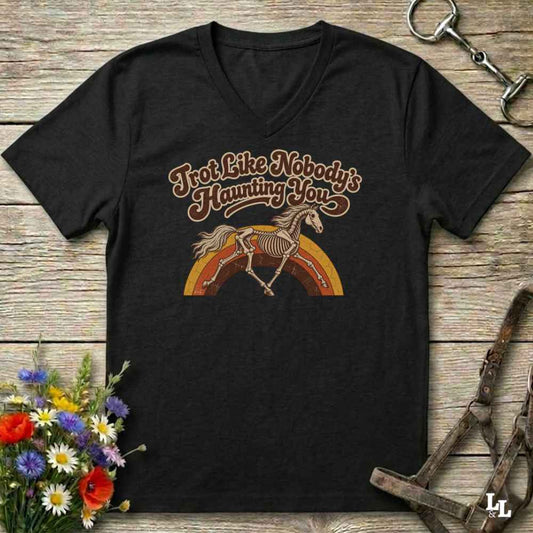 A black V-neck t-shirt with a retro graphic of a horse skeleton over a rainbow and the text 'I'm Not Like Nobody's Haunting You', displayed on a rustic wooden background with equestrian accessories and flowers.
