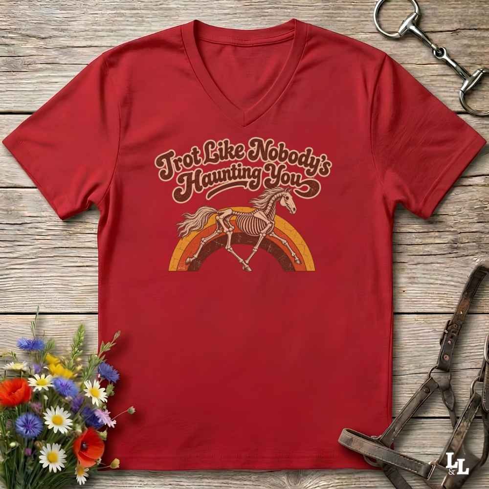 A red V-neck t-shirt laid flat on a rustic wooden surface, featuring a retro-style graphic of a horse skeleton under the text 'Trot Like Nobody's Haunting You' in brown and orange. The shirt is styled with equestrian accessories (a halter and bit) and a small bouquet of wildflowers.