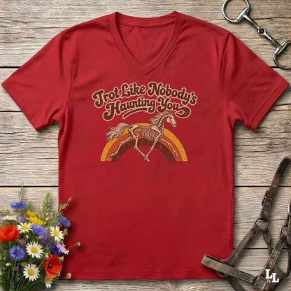 A red V-neck t-shirt laid flat on a rustic wooden surface, featuring a retro-style graphic of a horse skeleton under the text 'Trot Like Nobody's Haunting You' in brown and orange. The shirt is styled with equestrian accessories (a halter and bit) and a small bouquet of wildflowers.