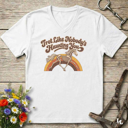 A white V-neck t-shirt with a retro-style graphic of a skeleton horse running over a rainbow, accompanied by the text 'Trot Like Nobody's Haunting You'. The shirt is displayed flat on a rustic wooden background with equestrian accessories and flowers.