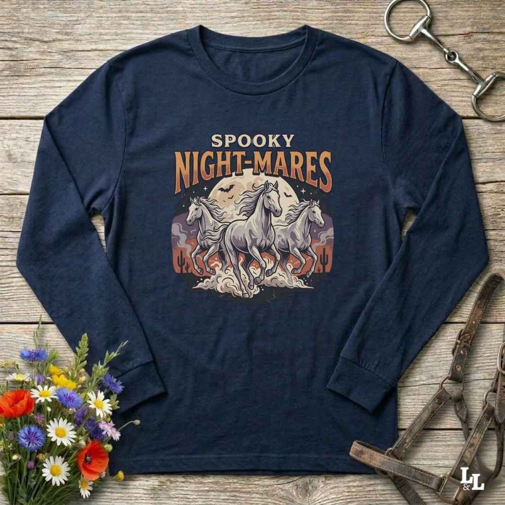 A navy blue long-sleeve t-shirt with a 'Spooky Night-Mares' graphic featuring three white horses and orange flames, laid flat on a rustic wooden background with decorative flowers and horse tack.