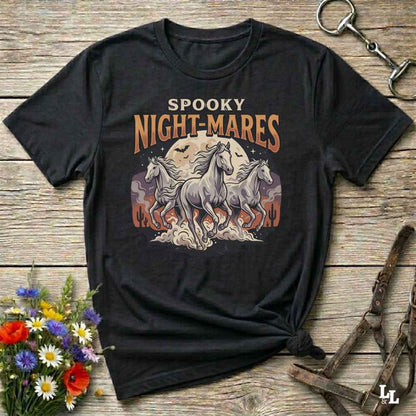 A black graphic t-shirt laid flat on a rustic wooden background, featuring a 'Spooky Night-Mares' design with three white horses in front of a full moon. The shirt is styled with a knot at the bottom hem, surrounded by decorative props including a small bouquet of wildflowers, a horse bit, and a leather bridle.