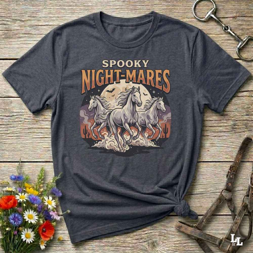 A dark heather gray graphic t-shirt featuring the text 'SPOOKY NIGHT-MARES' above an illustration of two white horses in front of a full moon. The shirt is styled in a flat-lay on a rustic wooden background with decorative flowers and horse tack.