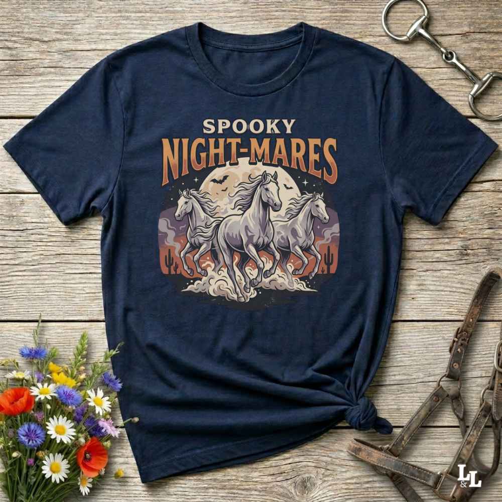 A navy blue graphic t-shirt laid flat on a rustic wooden surface, styled with equestrian accessories and a small bouquet of wildflowers. The shirt features a graphic print of three white horses in front of a full moon with the text 'SPOOKY NIGHT-MARES'.