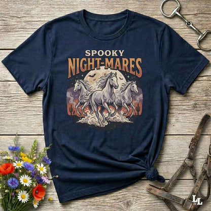 A navy blue graphic t-shirt laid flat on a rustic wooden surface, styled with equestrian accessories and a small bouquet of wildflowers. The shirt features a graphic print of three white horses in front of a full moon with the text 'SPOOKY NIGHT-MARES'.