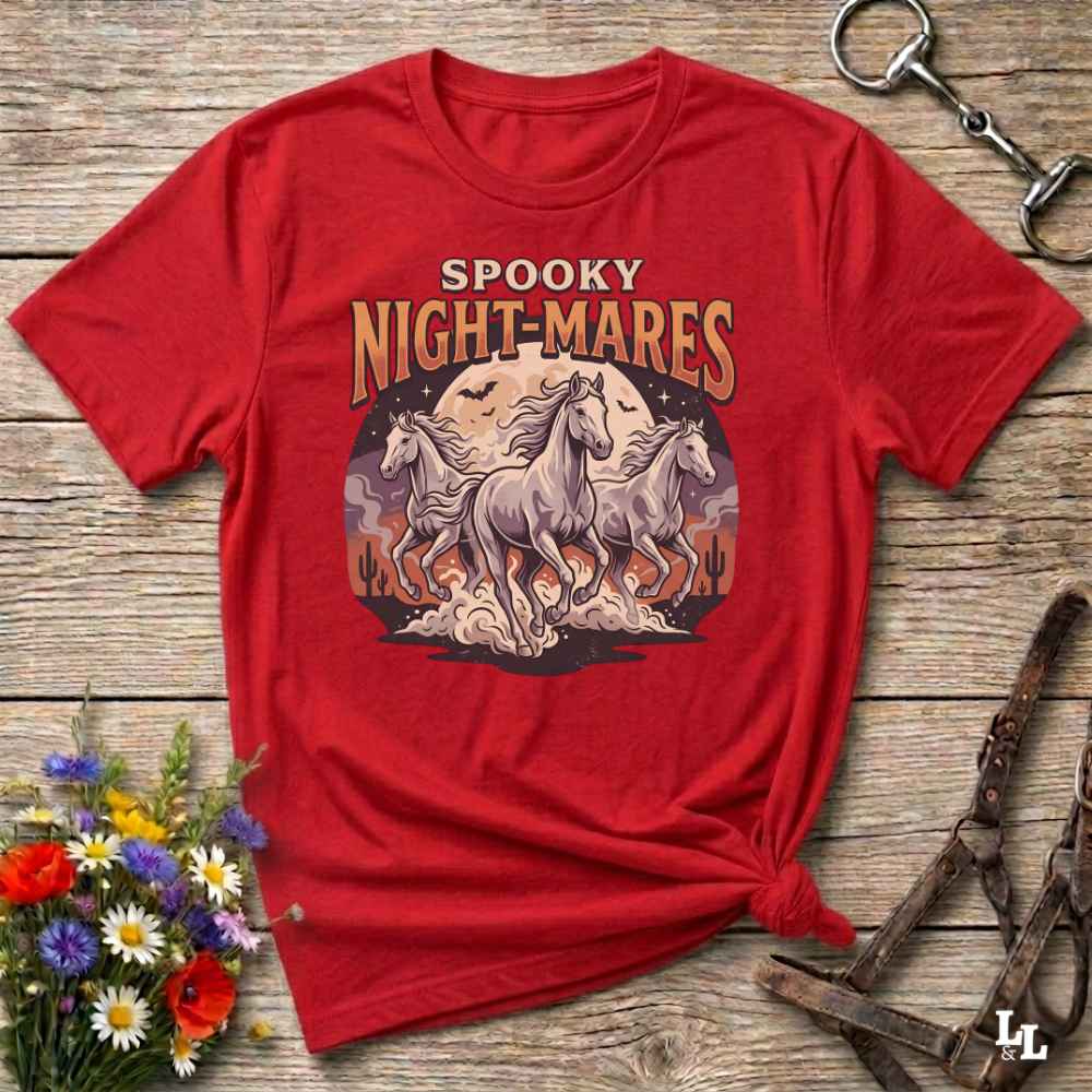 A red short-sleeve graphic T-shirt laid flat on a rustic wooden surface, styled with flowers, a horse bit, and a leather bridle. The shirt features a large vintage-style print reading 'SPOOKY NIGHT-MARES' above three white horses in front of a full moon and cacti.