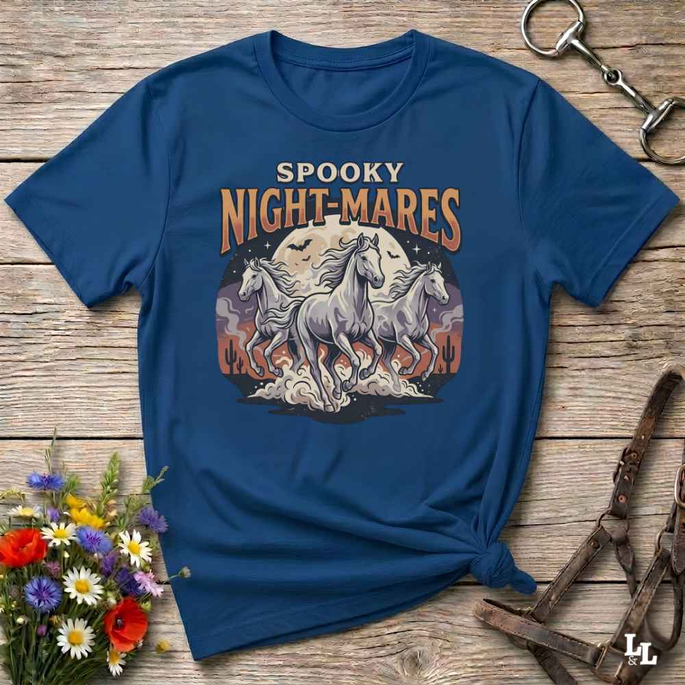 A navy blue graphic t-shirt laid flat on a rustic wooden surface, styled with a small bouquet of wildflowers, a horse bit, and a leather halter. The t-shirt features a graphic print of three white horses running in front of a full moon with the text 'SPOOKY NIGHT-MARES'.