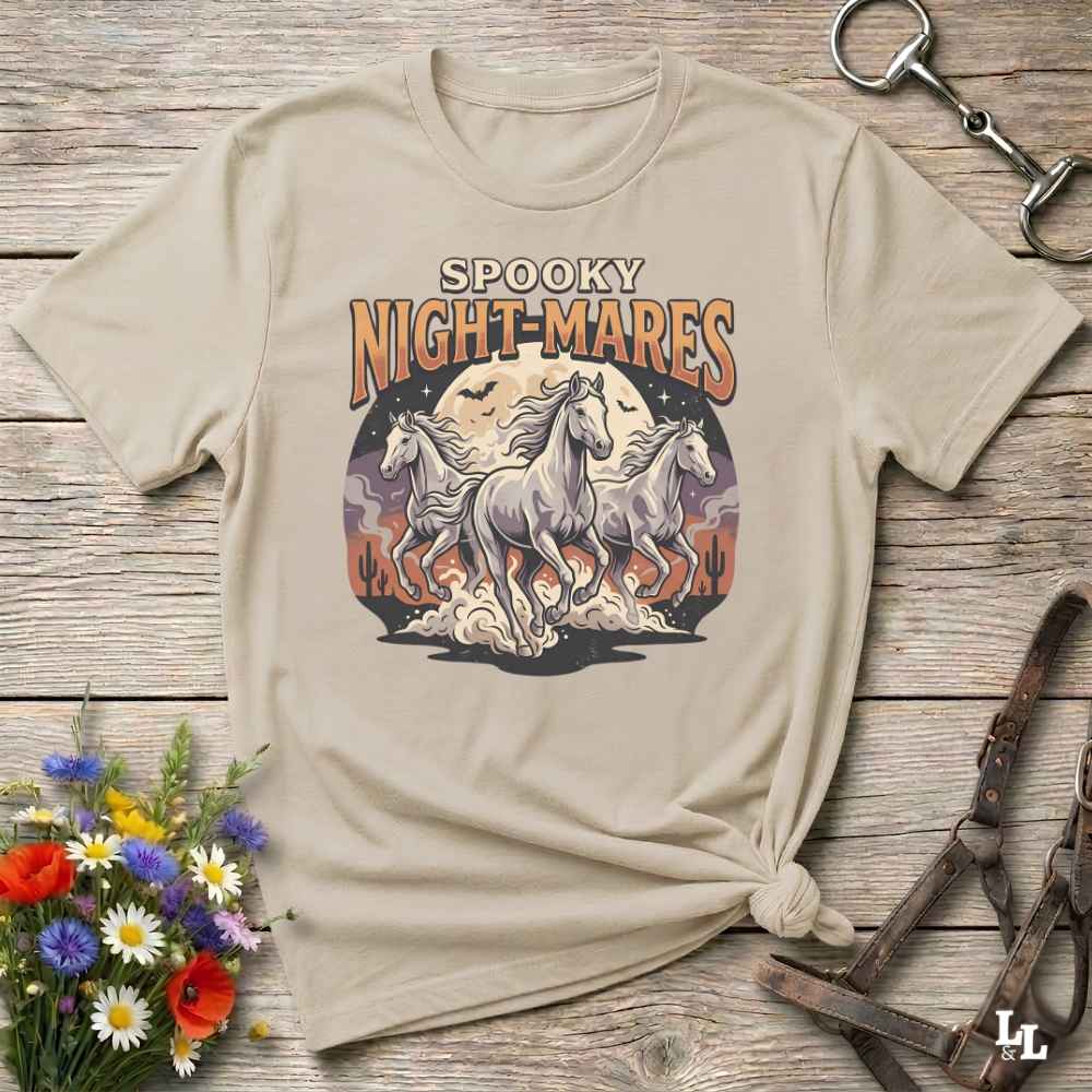 A beige graphic t-shirt with a 'Spooky Night-Mares' design featuring three white horses in front of a full moon, styled on a rustic wooden background with flowers and horse tack.