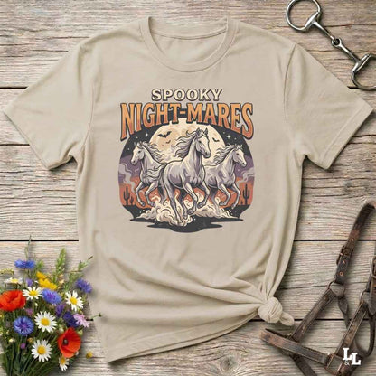 A beige graphic t-shirt with a 'Spooky Night-Mares' design featuring three white horses in front of a full moon, styled on a rustic wooden background with flowers and horse tack.