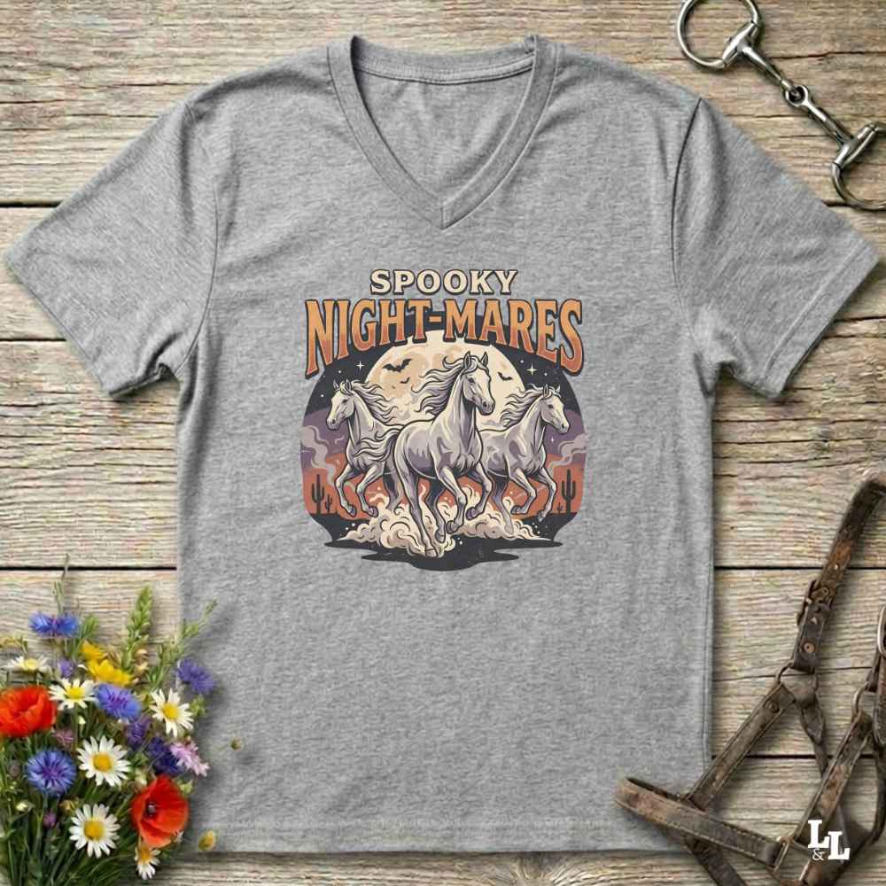A heather gray V-neck t-shirt with a 'Spooky Night-Mares' graphic print featuring three white horses in front of a full moon. The shirt is displayed flat on a rustic wooden background with decorative equestrian and floral props.