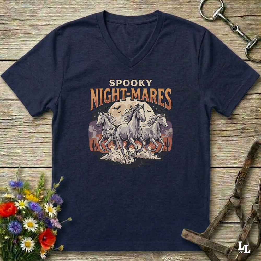 A navy blue V-neck t-shirt with a graphic print of three white horses in front of a full moon and the text 'SPOOKY NIGHT-MARES'. The shirt is displayed flat on a rustic wooden background with equestrian tack and a flower arrangement.
