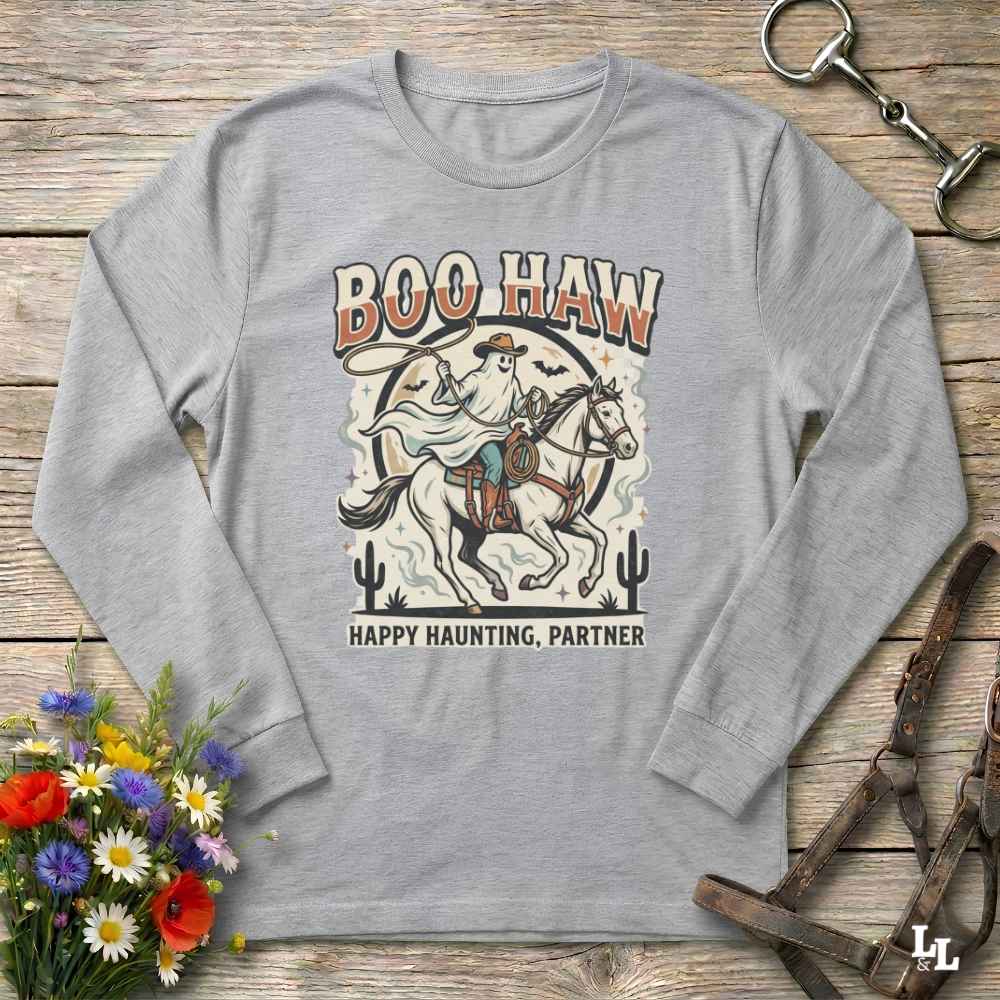 A light gray long-sleeve t-shirt with a vintage-style Halloween graphic showing a cowboy riding a horse with the text 'BOO HAW' and 'HAPPY HAUNTING, PARTNER'. The shirt is displayed flat on a rustic wooden background with decorative props including a bouquet of flowers, a horse bit, and a leather bridle.