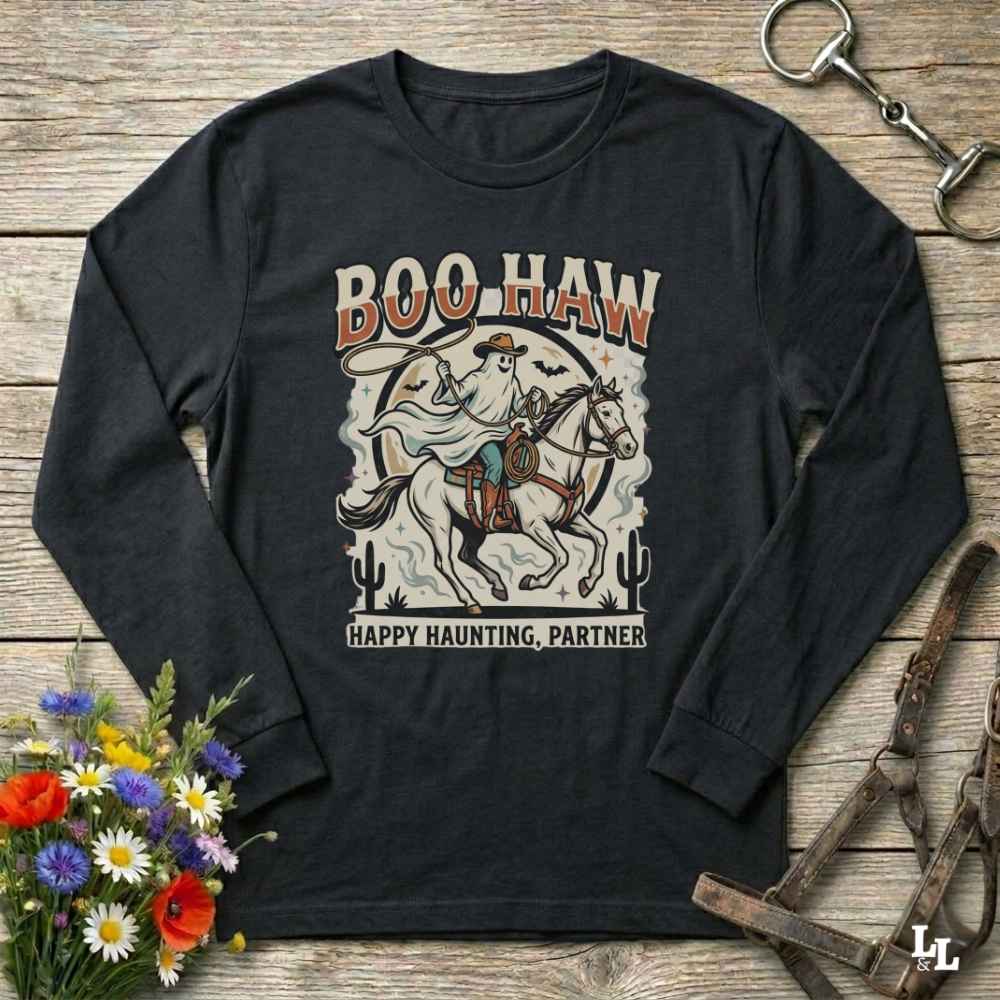 A black long-sleeve graphic t-shirt laid flat on a rustic wooden background, styled with a bouquet of colorful wildflowers, a horse bridle, and a metal tack piece. The shirt features a retro western-style graphic of a ghostly rider on a horse with the text 'BOO HAW' and 'HAPPY HAUNTING, PARTNER'.
