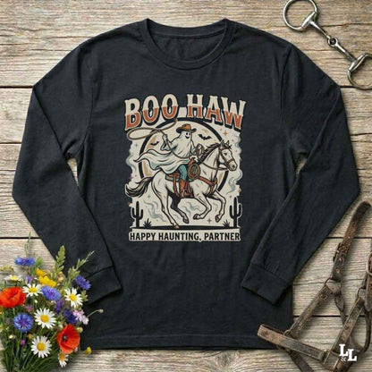 A black long-sleeve graphic t-shirt laid flat on a rustic wooden background, styled with a bouquet of colorful wildflowers, a horse bridle, and a metal tack piece. The shirt features a retro western-style graphic of a ghostly rider on a horse with the text 'BOO HAW' and 'HAPPY HAUNTING, PARTNER'.