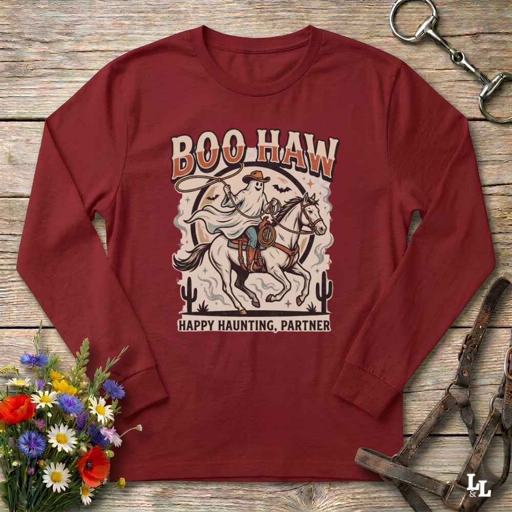 A long-sleeve crewneck t-shirt in maroon red, laid flat on a rustic wooden surface. The shirt features a vintage-style graphic of a cowboy riding a horse with the text 'BOO HAW' above and 'HAPPY HAUNTING, PARTNER' below.