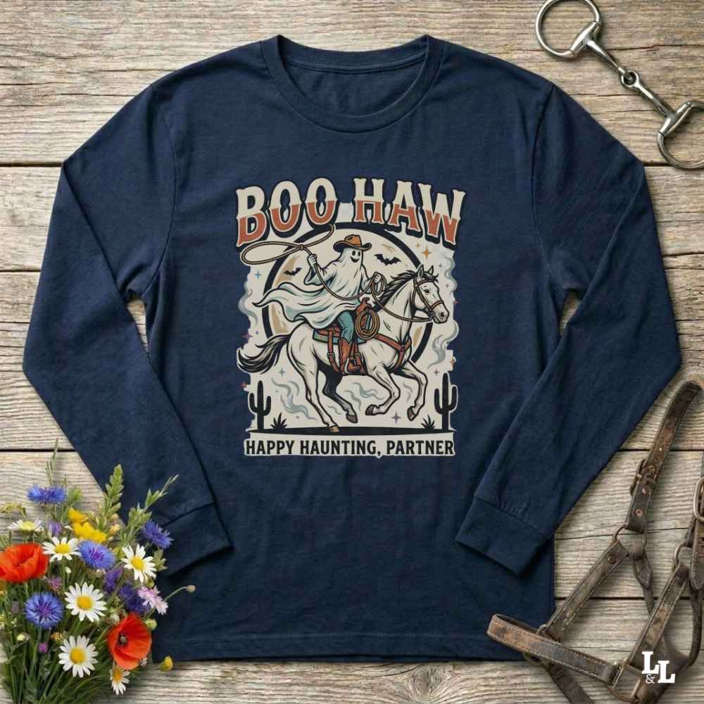 A navy blue long-sleeve graphic t-shirt laid flat on a rustic wooden background, surrounded by decorative props including a bouquet of flowers, a horse bit, and a leather bridle. The shirt features a western-themed graphic of a cowboy on a horse with the text 'BOO HAW' and 'HAPPY HAUNTING, PARTNER'.