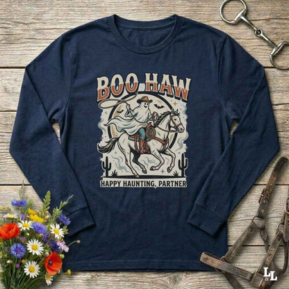 A navy blue long-sleeve graphic t-shirt laid flat on a rustic wooden background, surrounded by decorative props including a bouquet of flowers, a horse bit, and a leather bridle. The shirt features a western-themed graphic of a cowboy on a horse with the text 'BOO HAW' and 'HAPPY HAUNTING, PARTNER'.