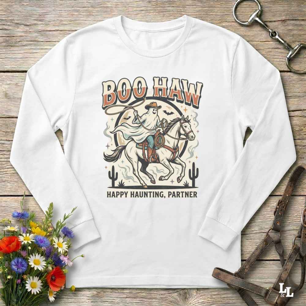 A white long-sleeve t-shirt with a western-themed 'Boo Haw Happy Haunting, Partner' graphic print, laid flat on a rustic wooden background with equestrian and floral props.