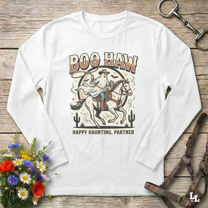 A white long-sleeve t-shirt with a western-themed 'Boo Haw Happy Haunting, Partner' graphic print, laid flat on a rustic wooden background with equestrian and floral props.